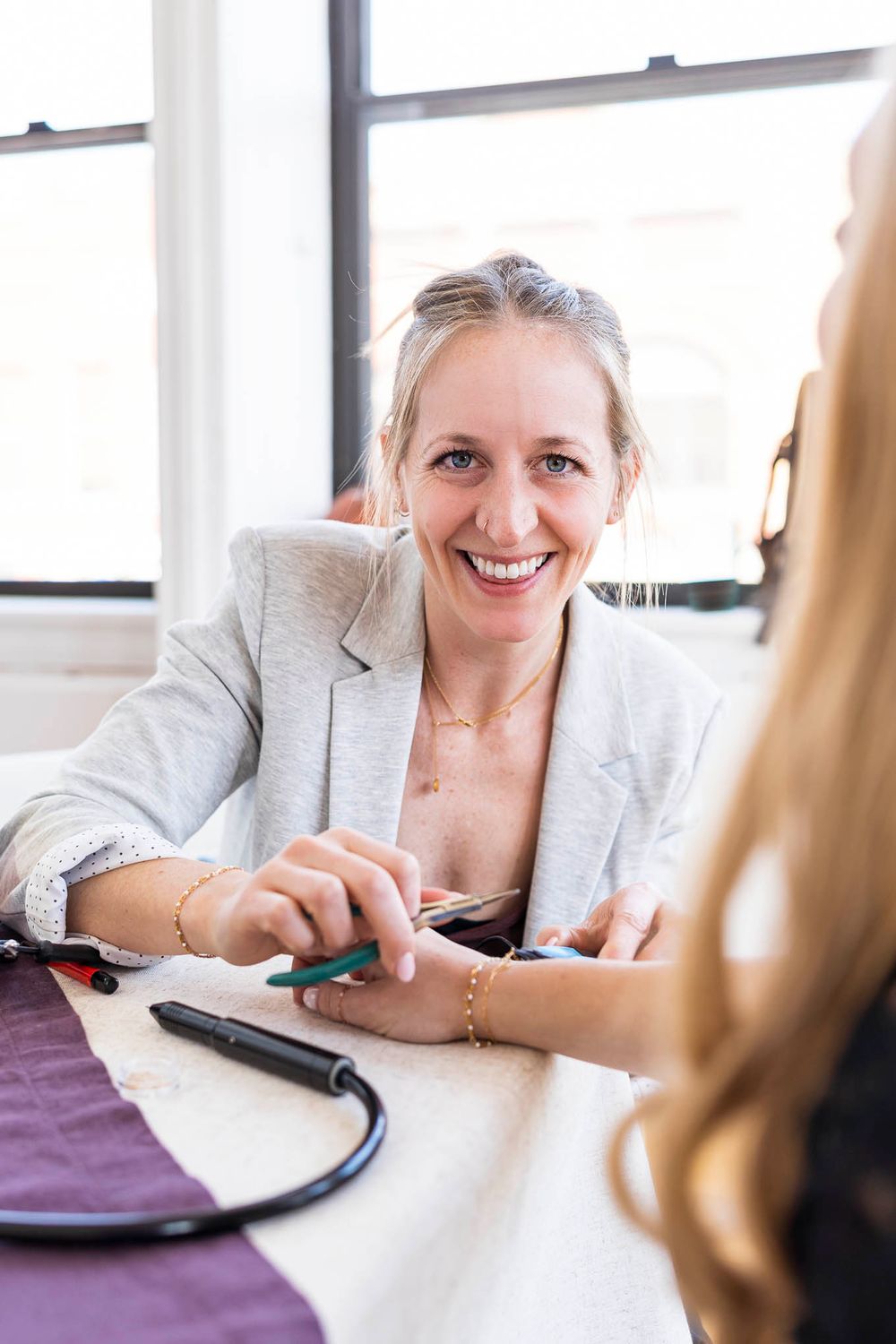 Person in light blazer smiling while working at table with jewelry supplies.