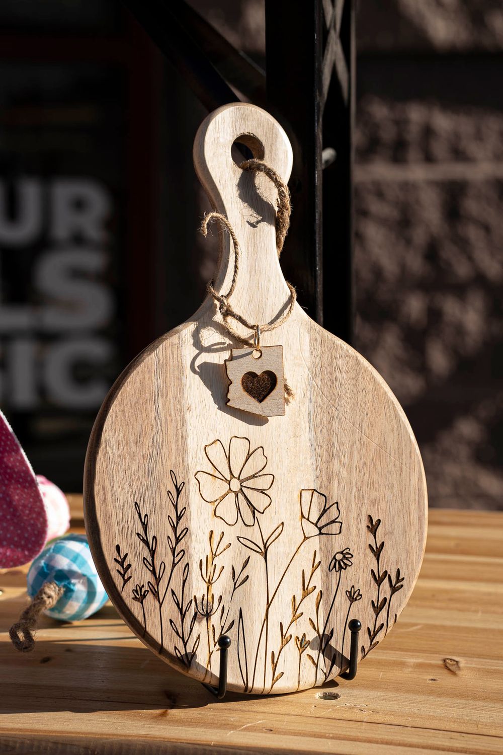 Wooden cutting board with floral design etched into surface and heart charm hanging from handle.