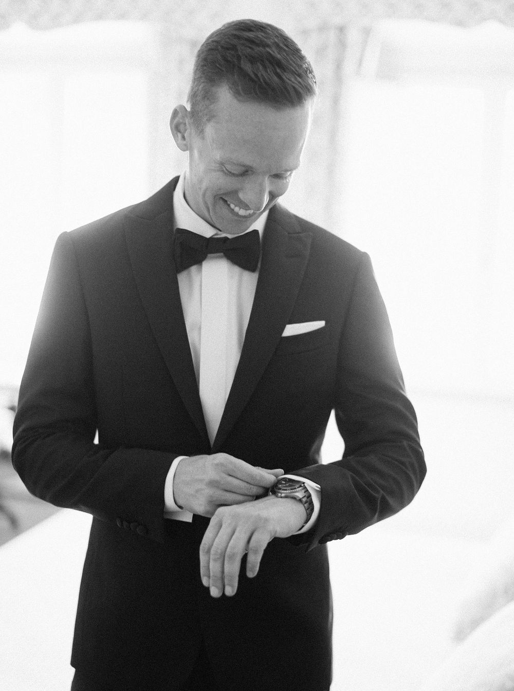 A well-dressed groom in a tuxedo carefully adjusts his watch, exuding elegance and sophistication