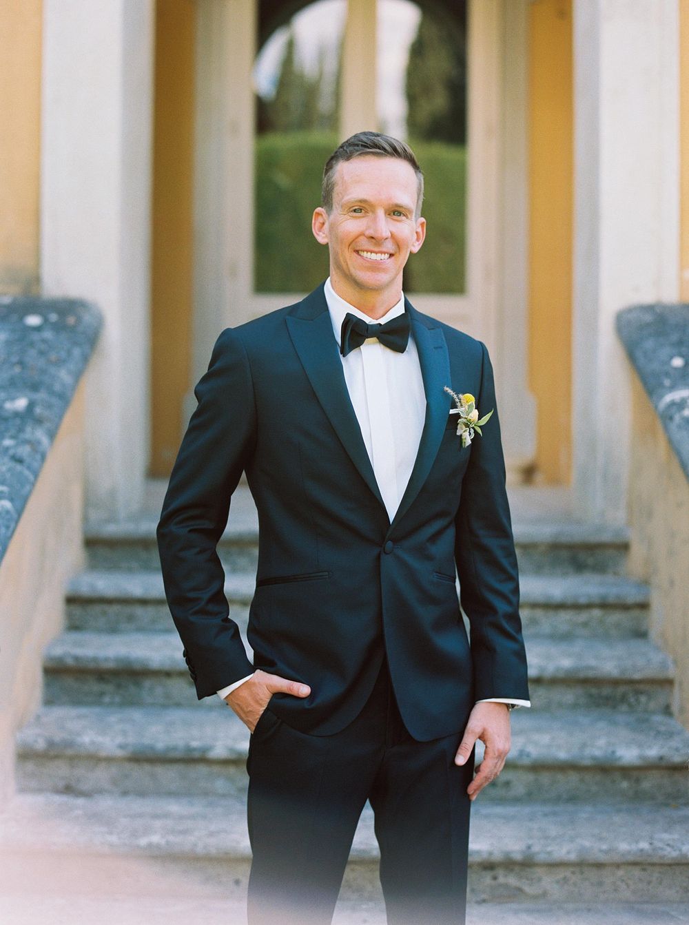An elegantly dressed groom poses for a photo in front of villa.