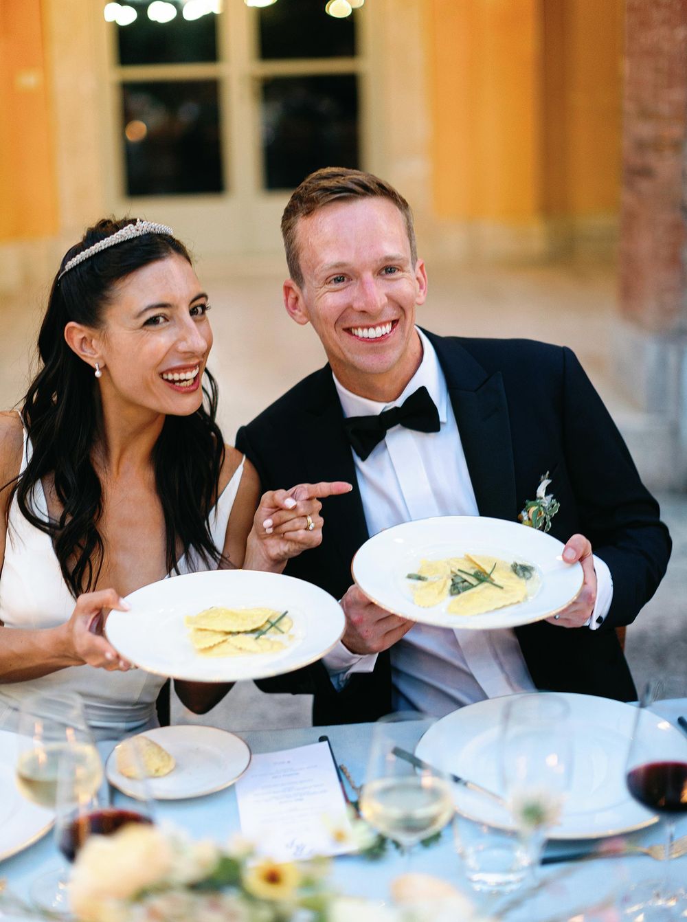 Bride and groom smiling, holding plates of food at a wedding in Tuscany.