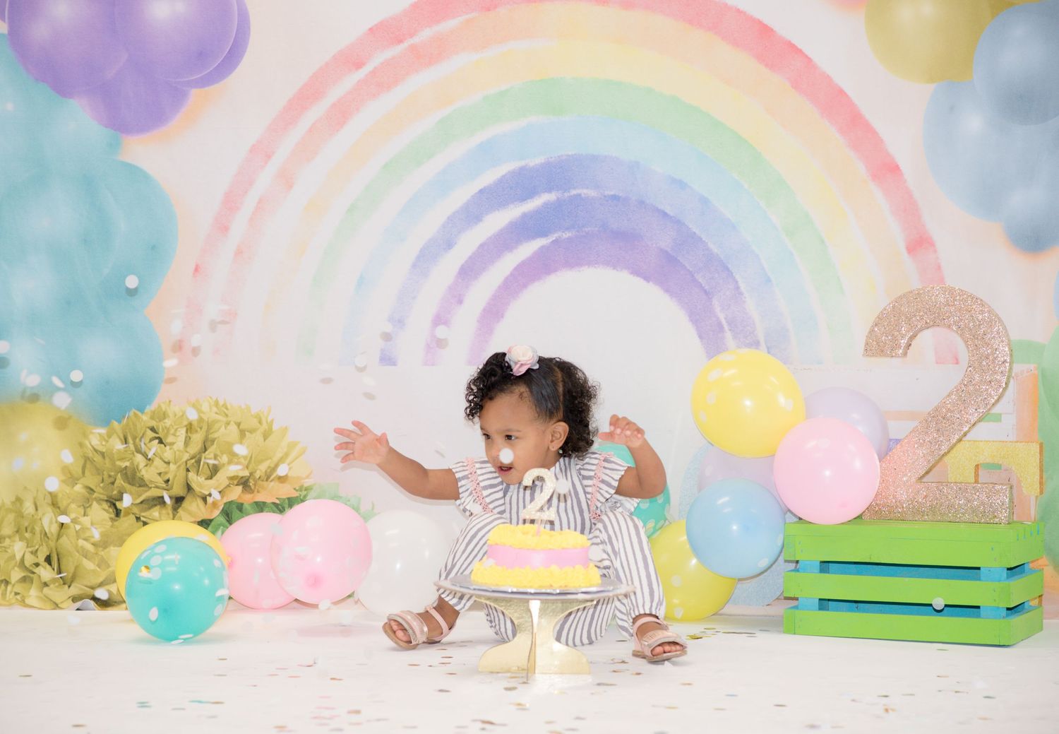 Rainbow themed birthday photoshoot with toddler playing amid colorful balloons.
