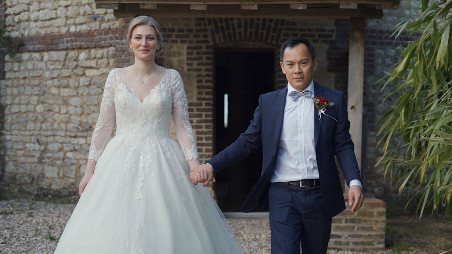 A bride in a lace wedding gown and groom in navy suit pose together against a brick wall backdrop.