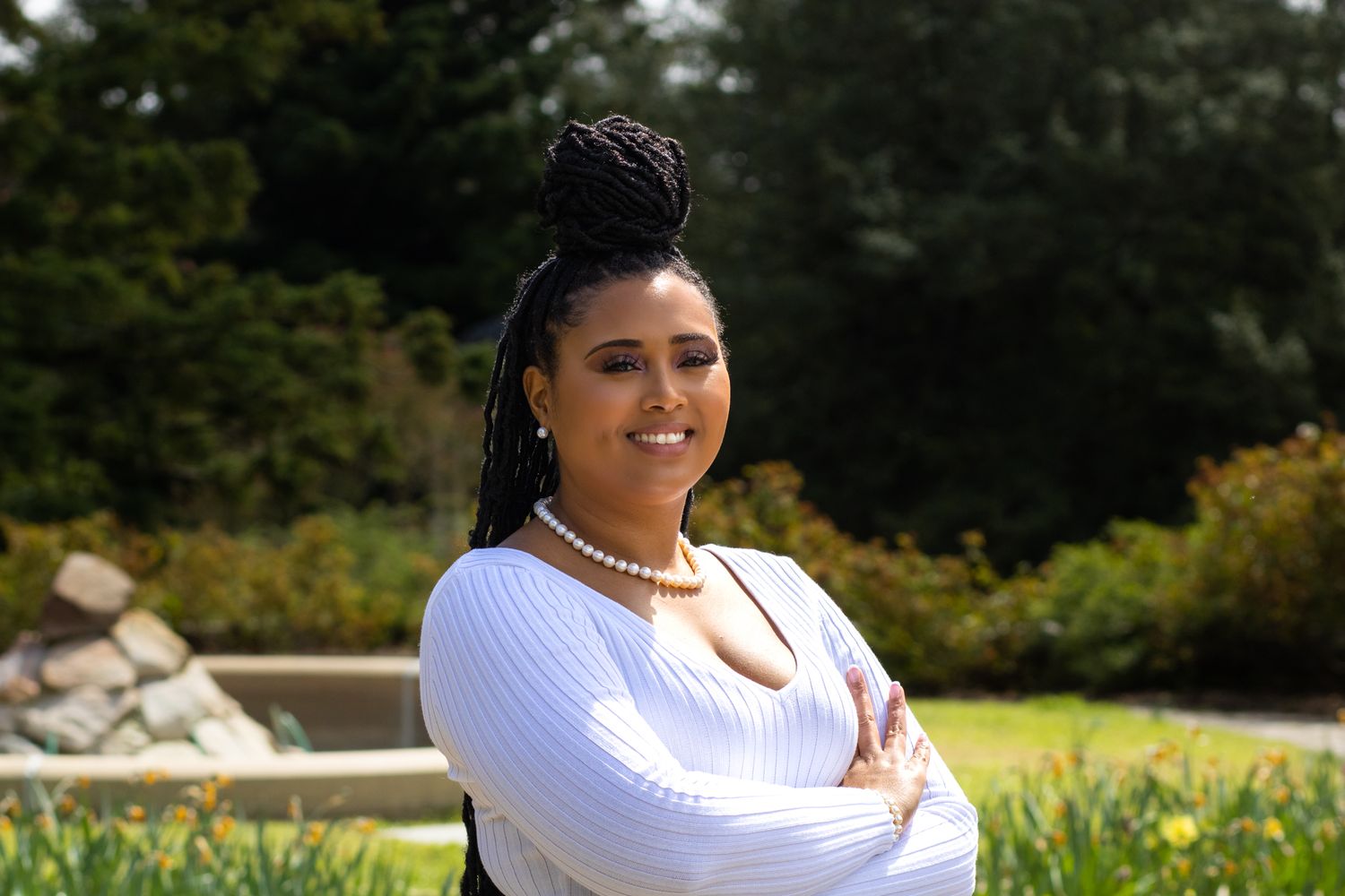 Smiling woman in white sweater with pearl necklace and braids posing in garden setting.