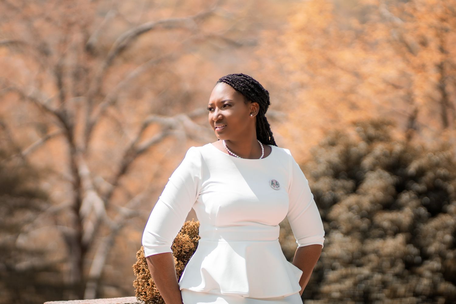 Woman in white peplum dress standing outdoors with autumn trees in background.