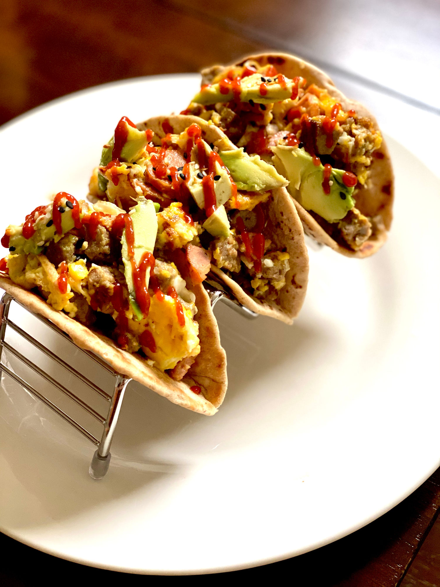 Three loaded breakfast tacos with scrambled eggs, avocado, and sriracha drizzle served in a metal taco holder on a white plate.