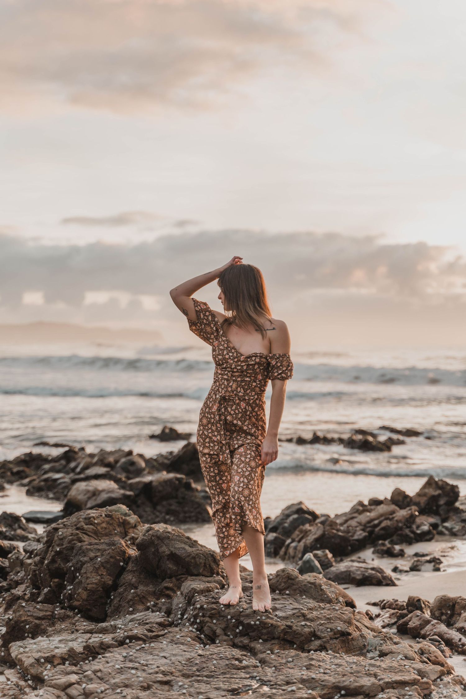Woman in leopard print dress stands on rocky beach at sunset.