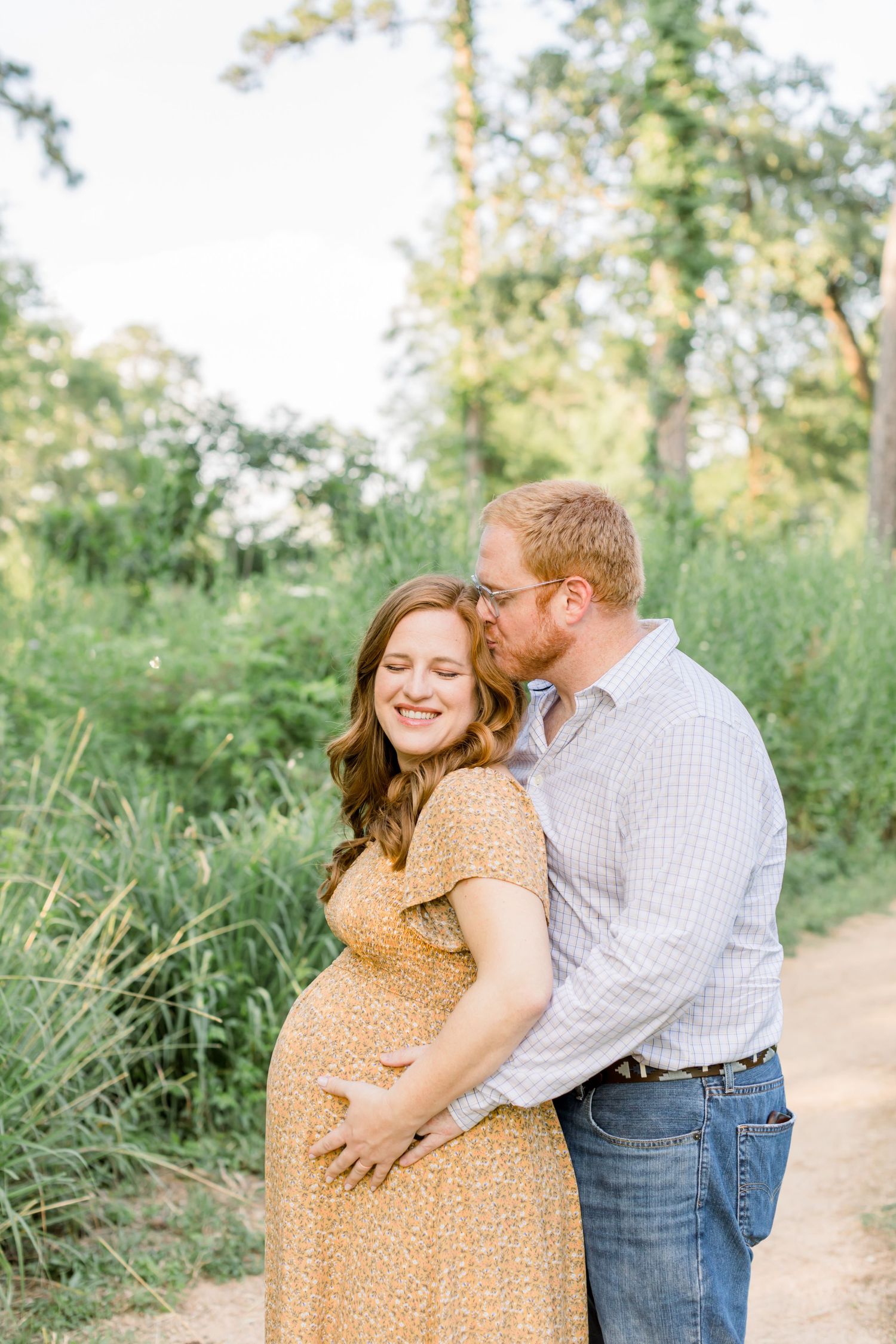 Couple shares tender moment on woodland path during pregnancy photoshoot.