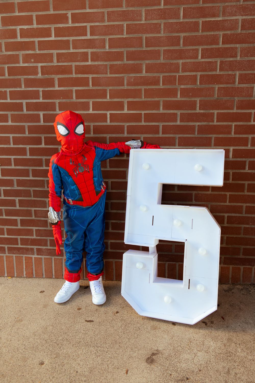 Person wearing Spider-Man costume poses next to large white number 5 against brick wall.