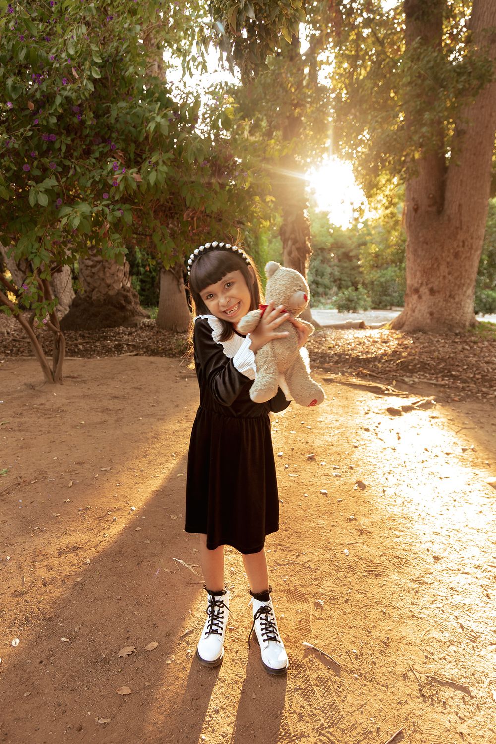 Someone in a black dress holds a stuffed animal while standing on a sunlit forest path.