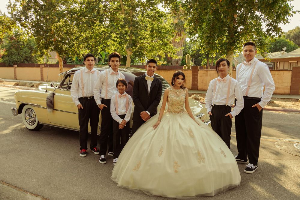 Wedding party poses with classic cars on a tree-lined street.