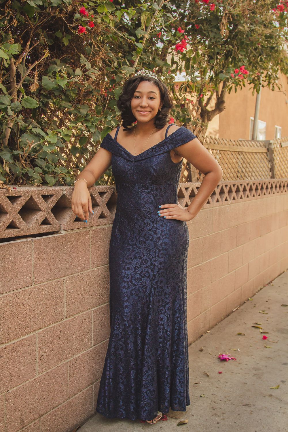 Full-length portrait showing off navy lace evening gown against decorative wall and red flowers.