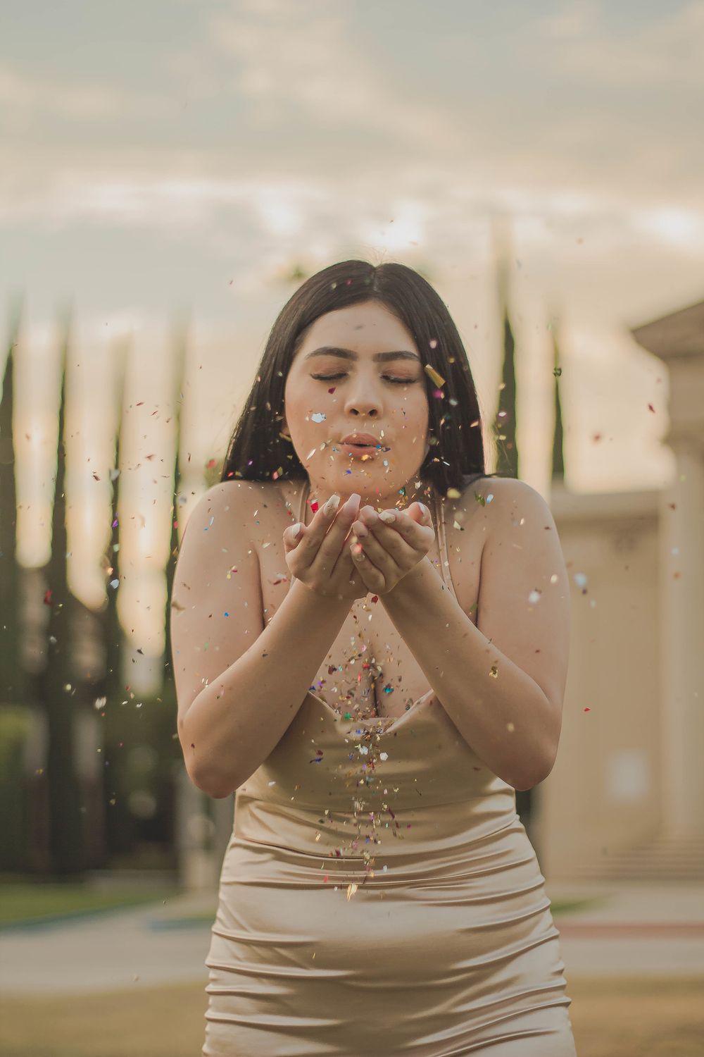 A person in a beige dress blows glitter while standing in soft outdoor lighting.