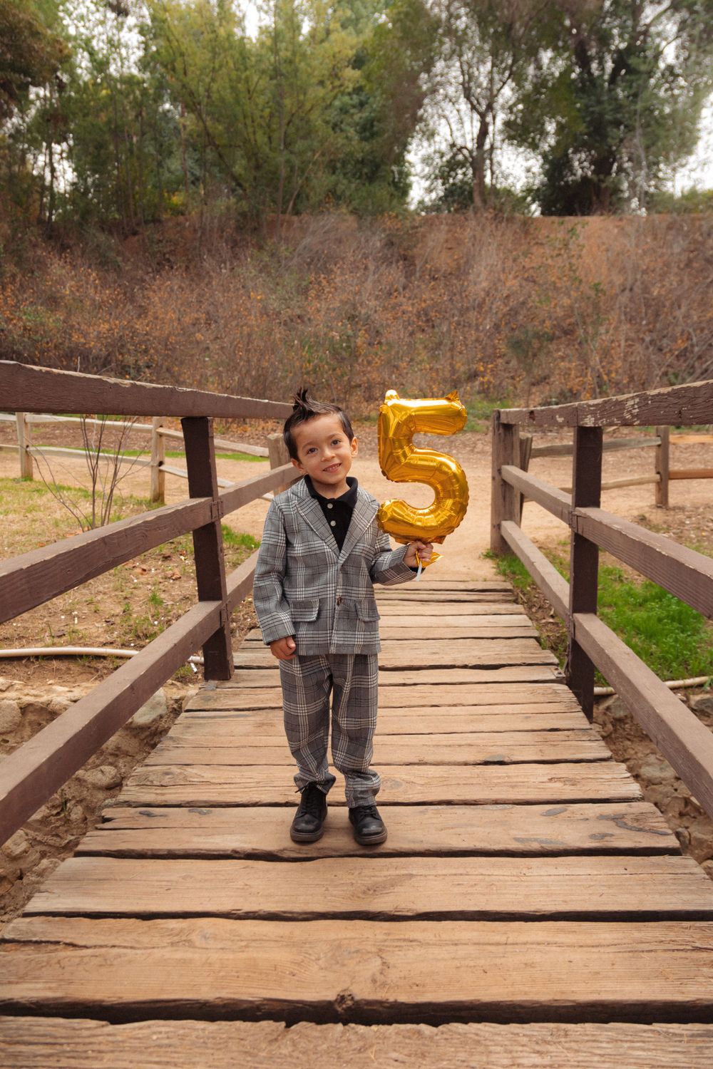 A child in a plaid suit holds a gold number 5 balloon while standing on a wooden bridge.