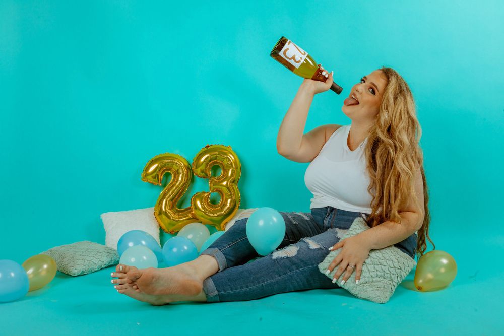Someone drinking from a bottle with golden 23 balloons and scattered glitter on turquoise background.