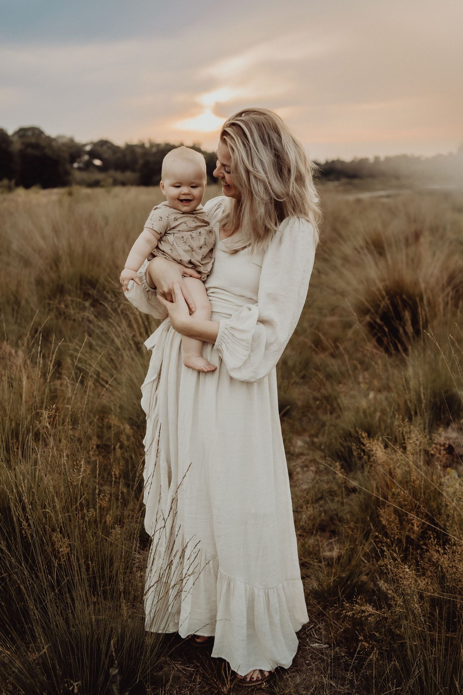 Mother and baby in flowing white dress stand in golden grassy field at sunset.