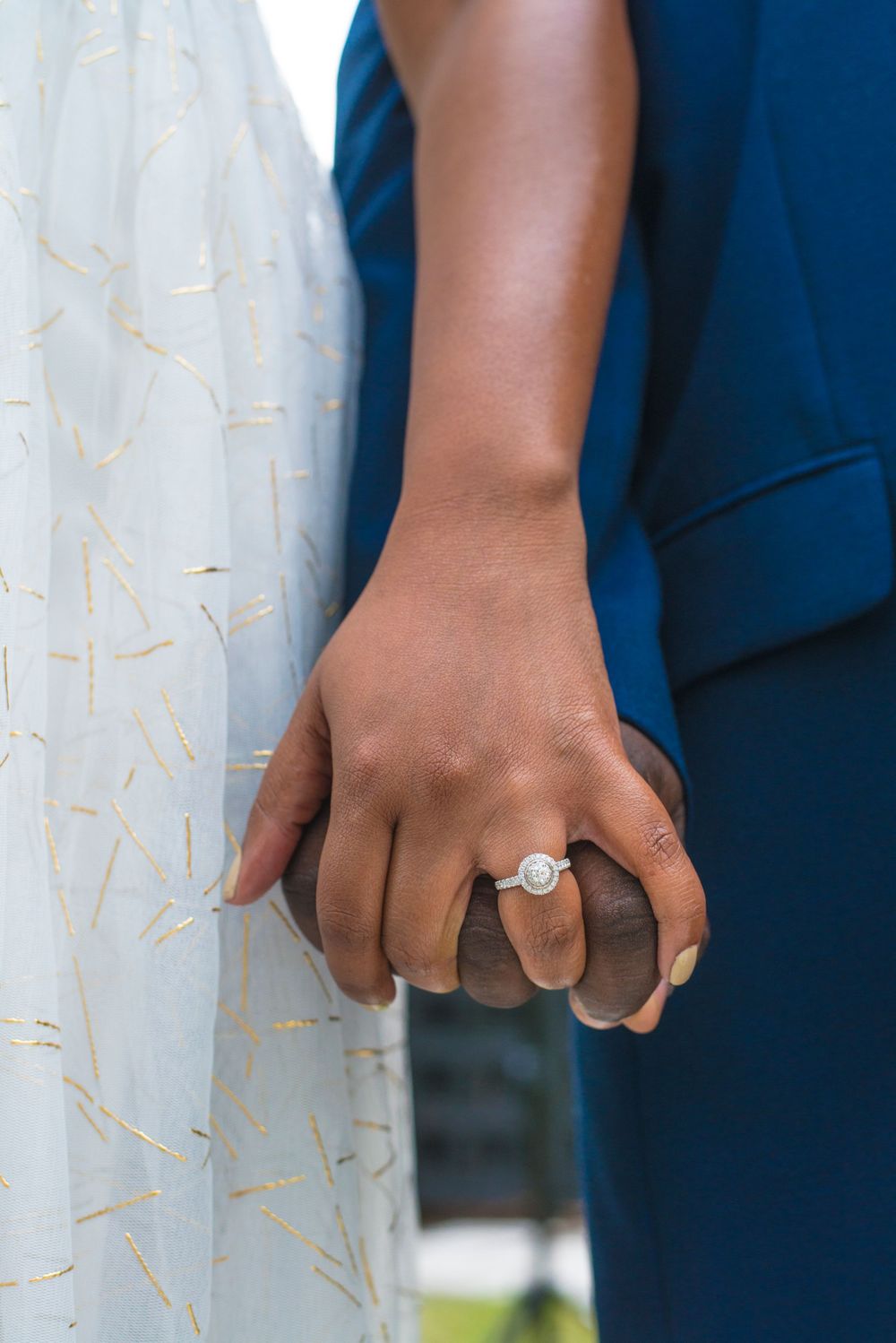 Close-up of interracial couple holding hands showing engagement ring against white dress.