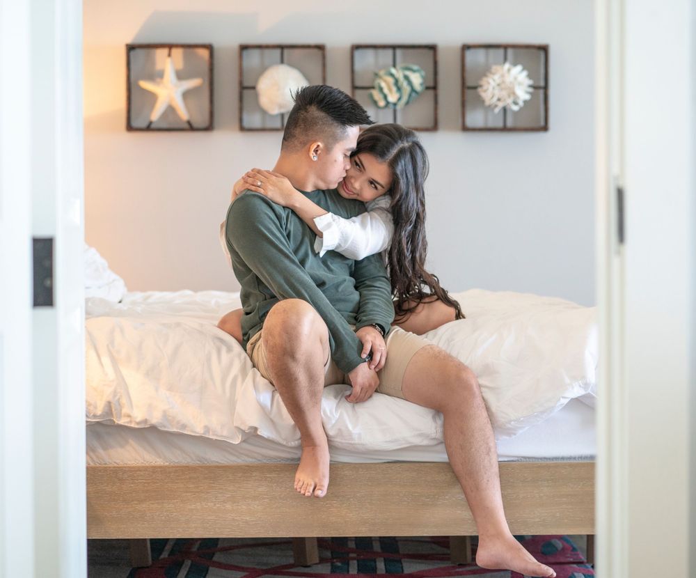 A romantic couple sharing an intimate moment while sitting on a bed with coastal artwork on the wall.