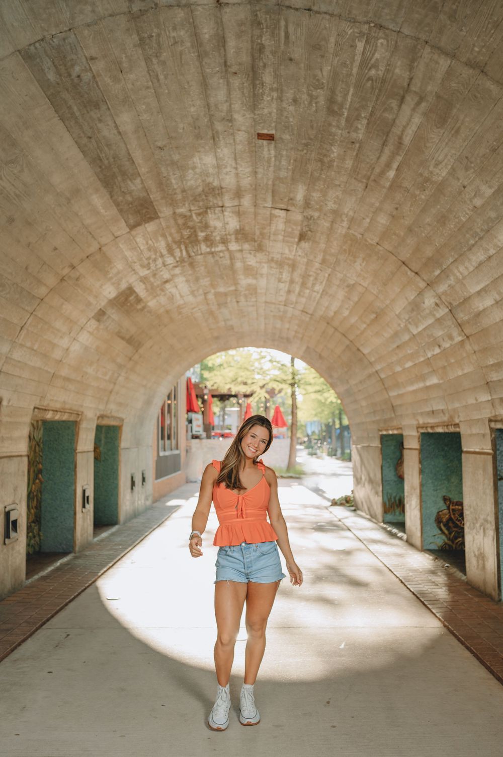 Person wearing orange top and denim shorts standing in arched brick tunnel walkway.