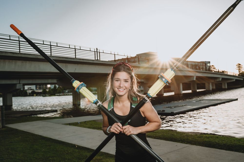 Athlete posing with crossed rowing oars at sunset near a bridge over water.