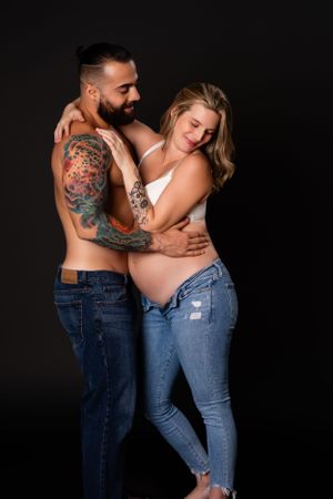 Couple in casual denim share tender moment during maternity photo session against black backdrop.