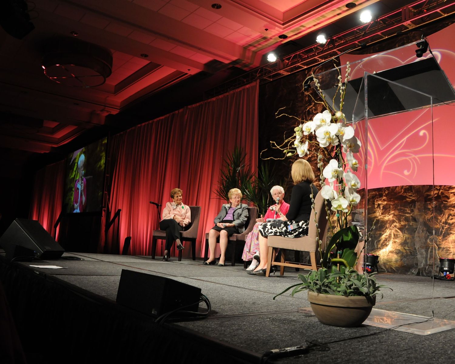 Panel discussion setup on stage with red curtains and decorative floral arrangements.
