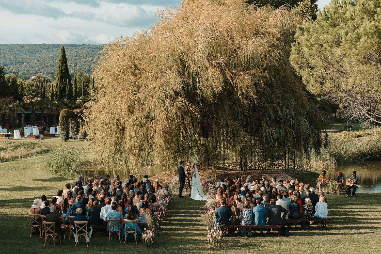 wedding photo of bride and groom under a tree canopy at the top of aisle during the ceremony in Provence, France.