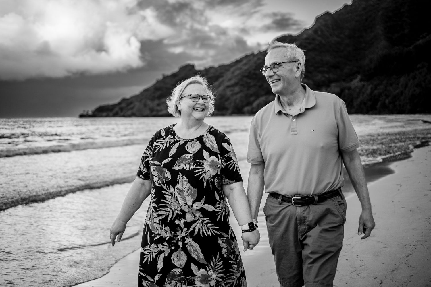 A couple walking together on a scenic beach with mountains and dramatic clouds.