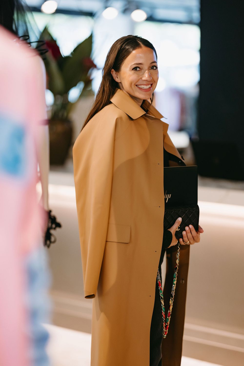 Professional wearing stylish camel coat holding laptop in modern office setting.