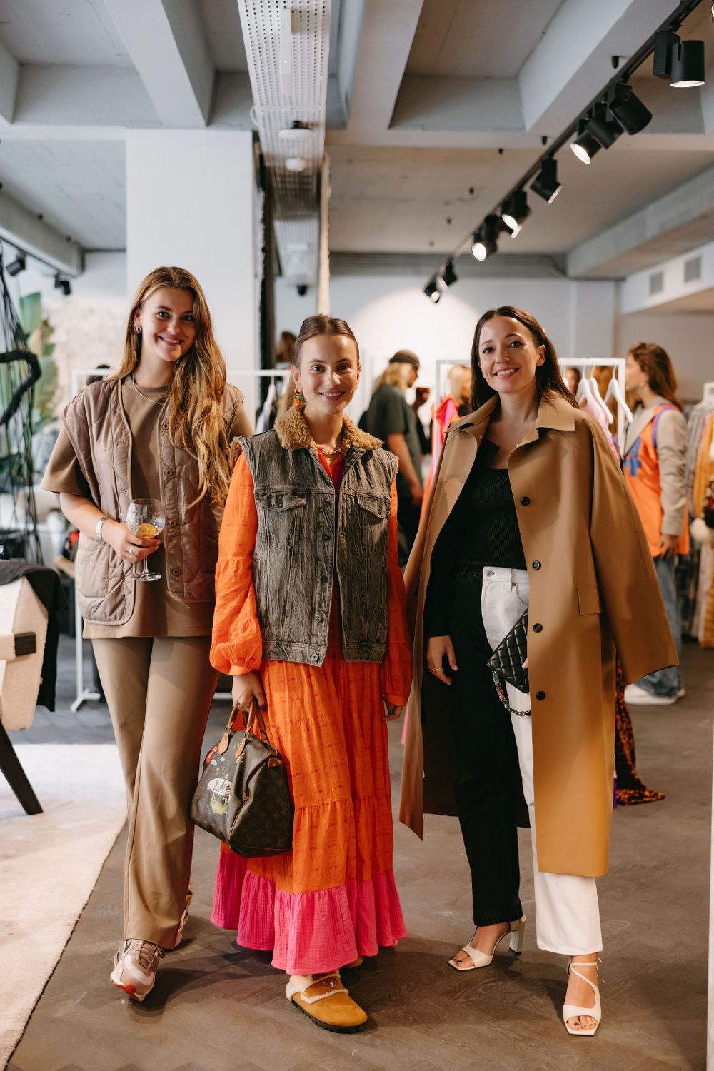 Three friends showcase contrasting fashion styles including a camel coat, orange dress, and neutral outfit.