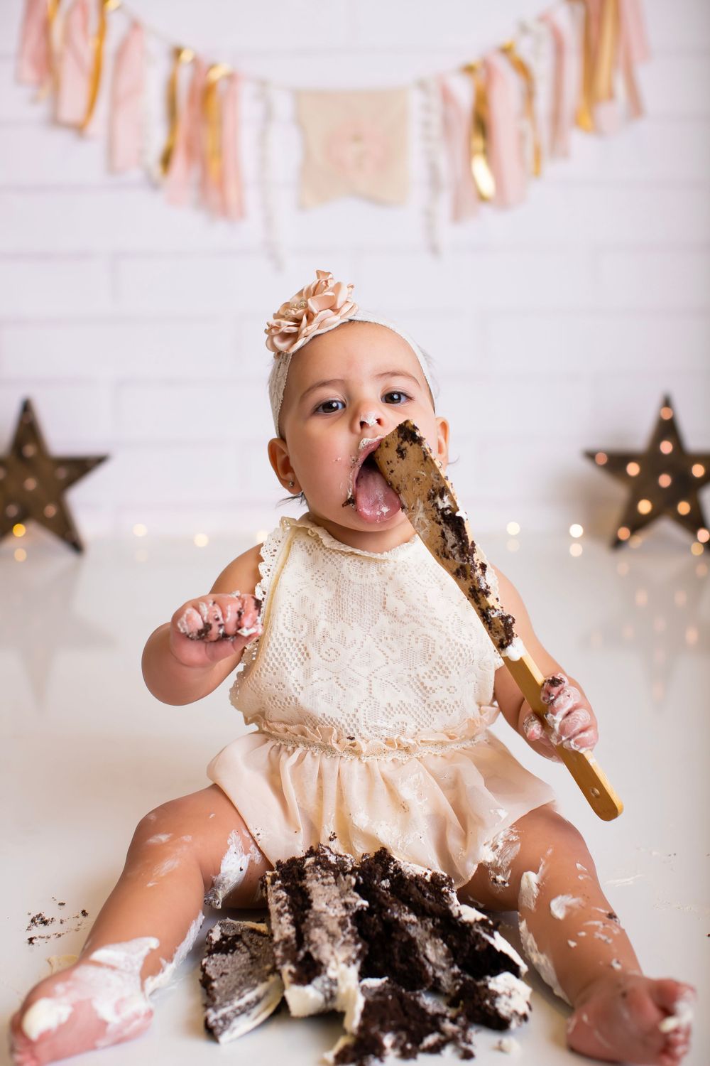 Baby in cream dress enjoying smash cake photoshoot with decorative stars and banner in background.