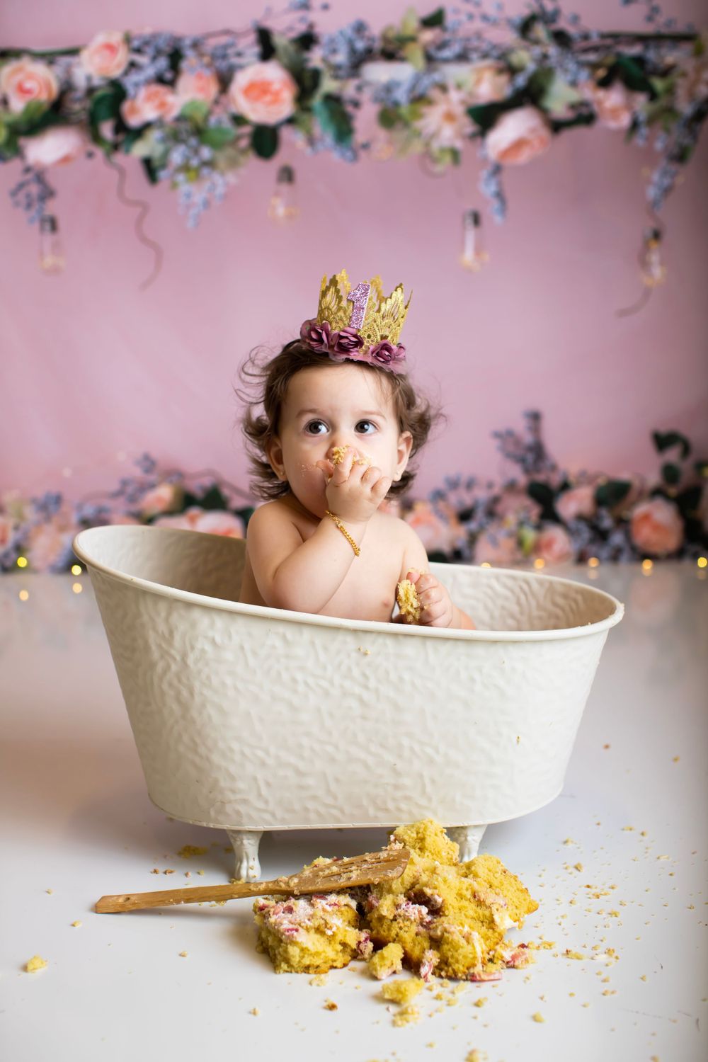 Baby in bathtub wearing gold crown during cake smash photography session with floral backdrop.