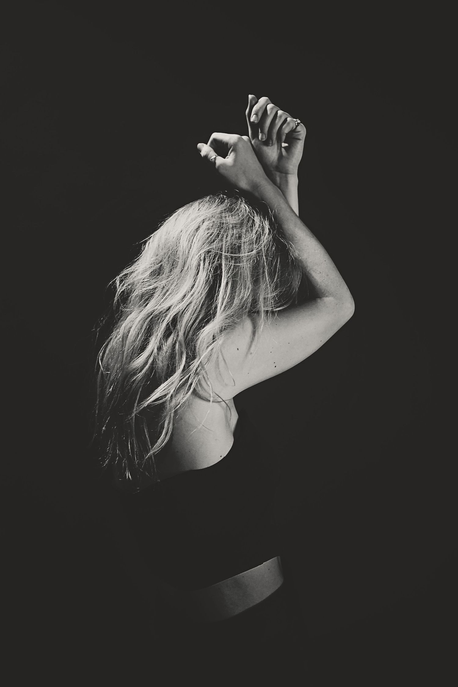 Artistic black and white portrait of woman with flowing blonde hair with raised arms against dark background.