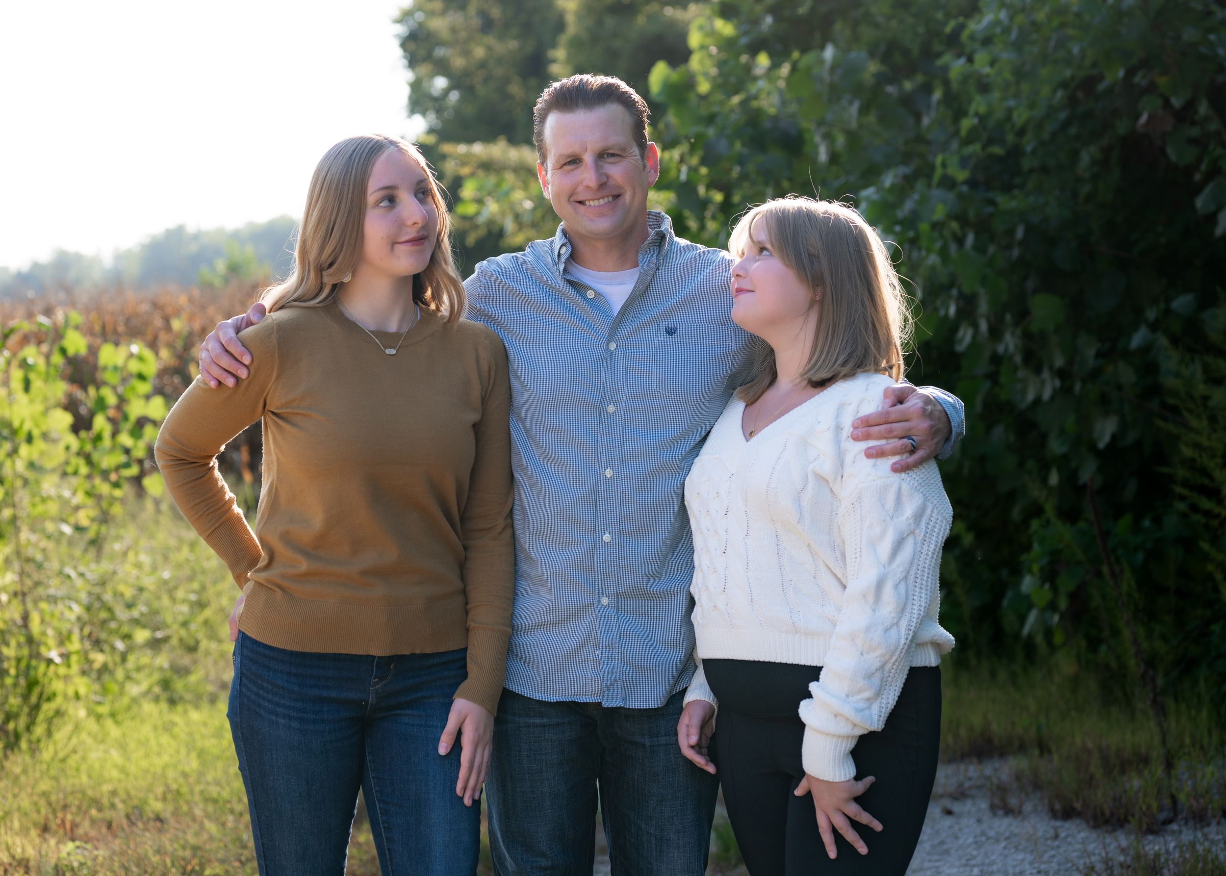 Three people share casual embrace while standing on rural path at sunset.