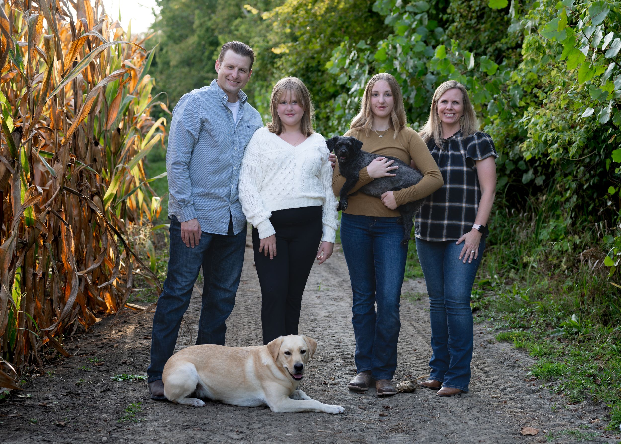 Family poses for a portrait on a path with their golden retriever beside a corn field.