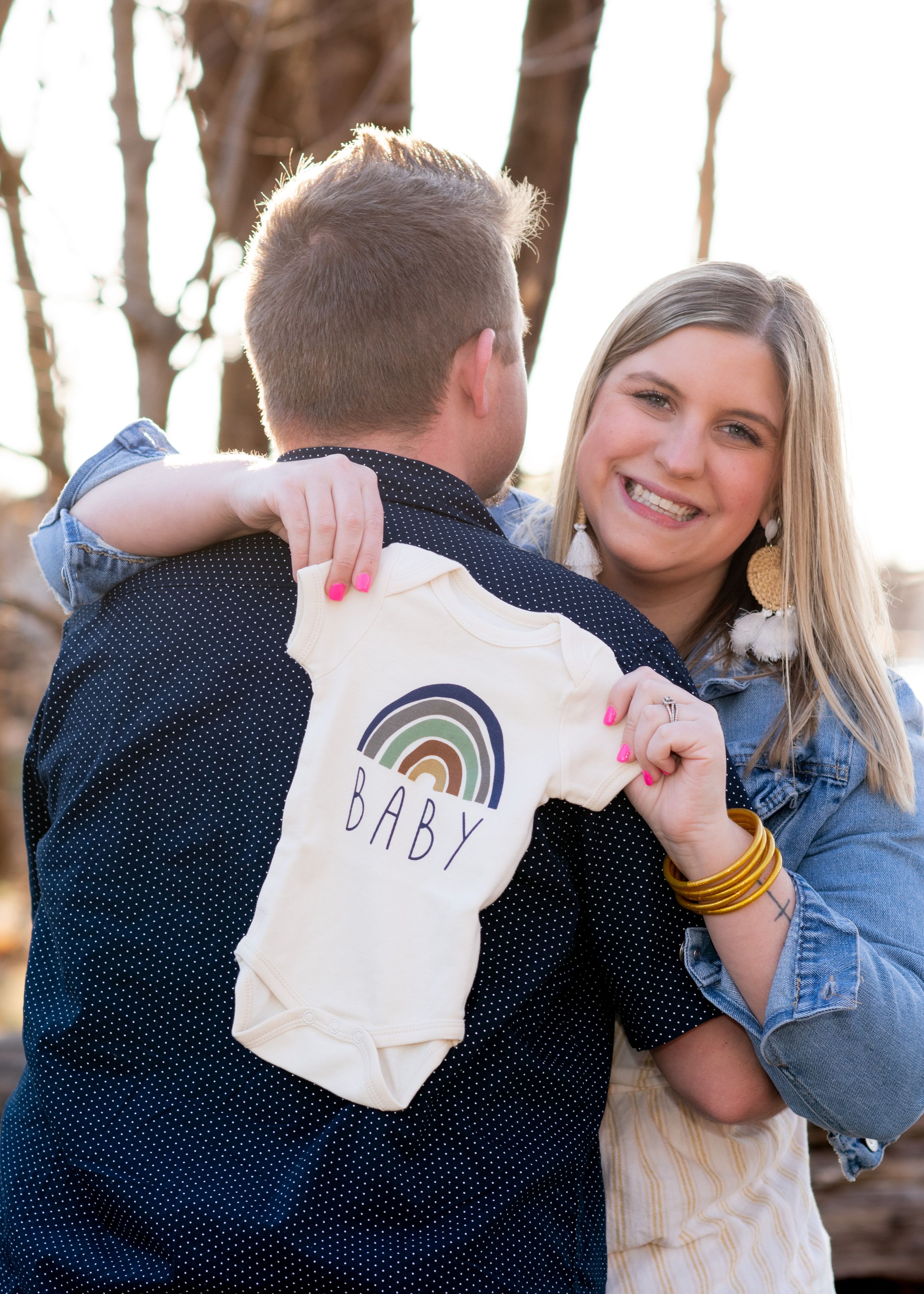 A couple displays a rainbow-decorated baby onesie outdoors in natural lighting.
