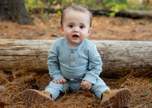 Baby in light blue sweater and pants sitting on fallen log in autumn forest setting.