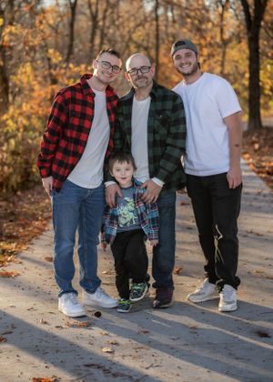 Four generations pose together on an autumn path lined with fallen leaves.