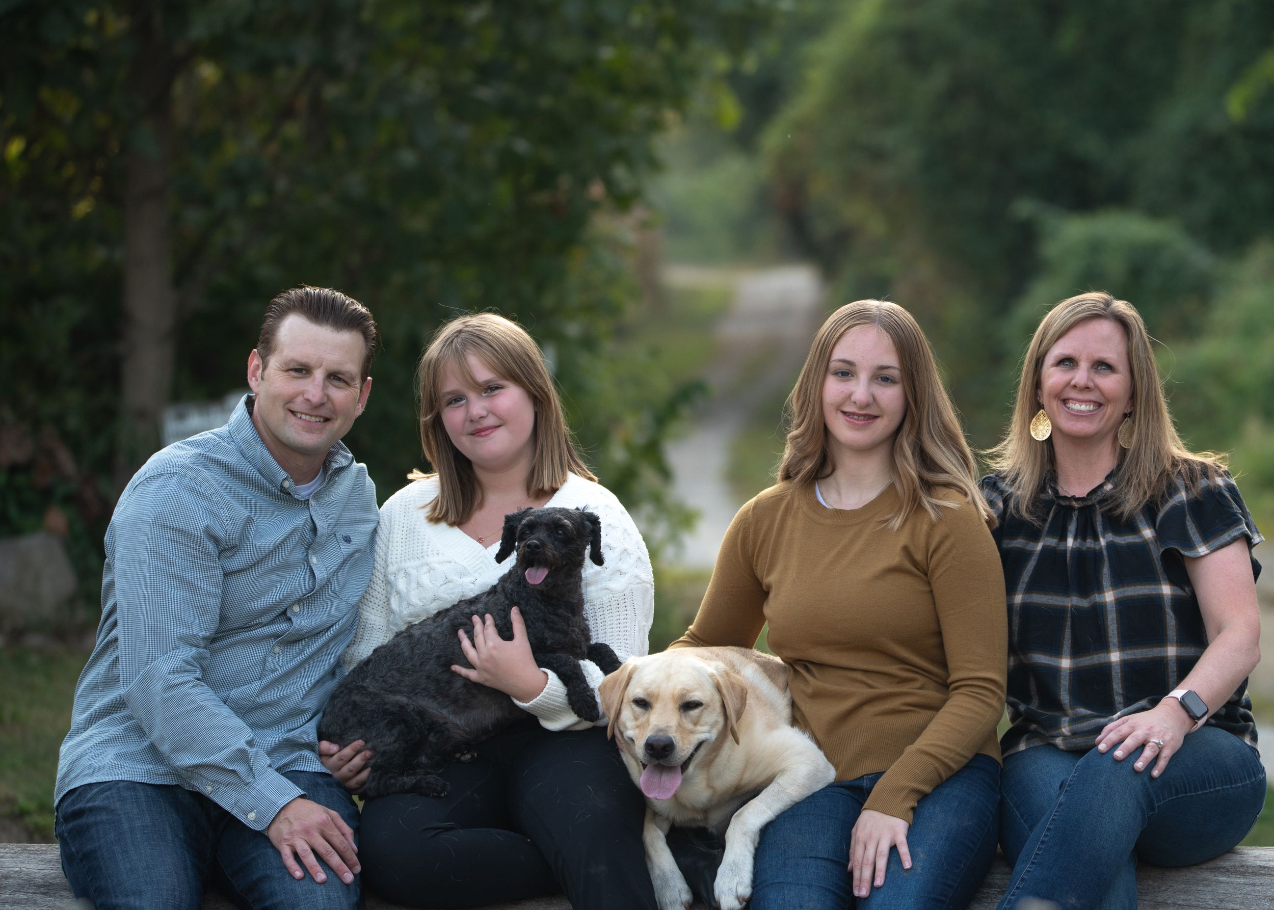 Family group sits outdoors with two dogs on sunny day.