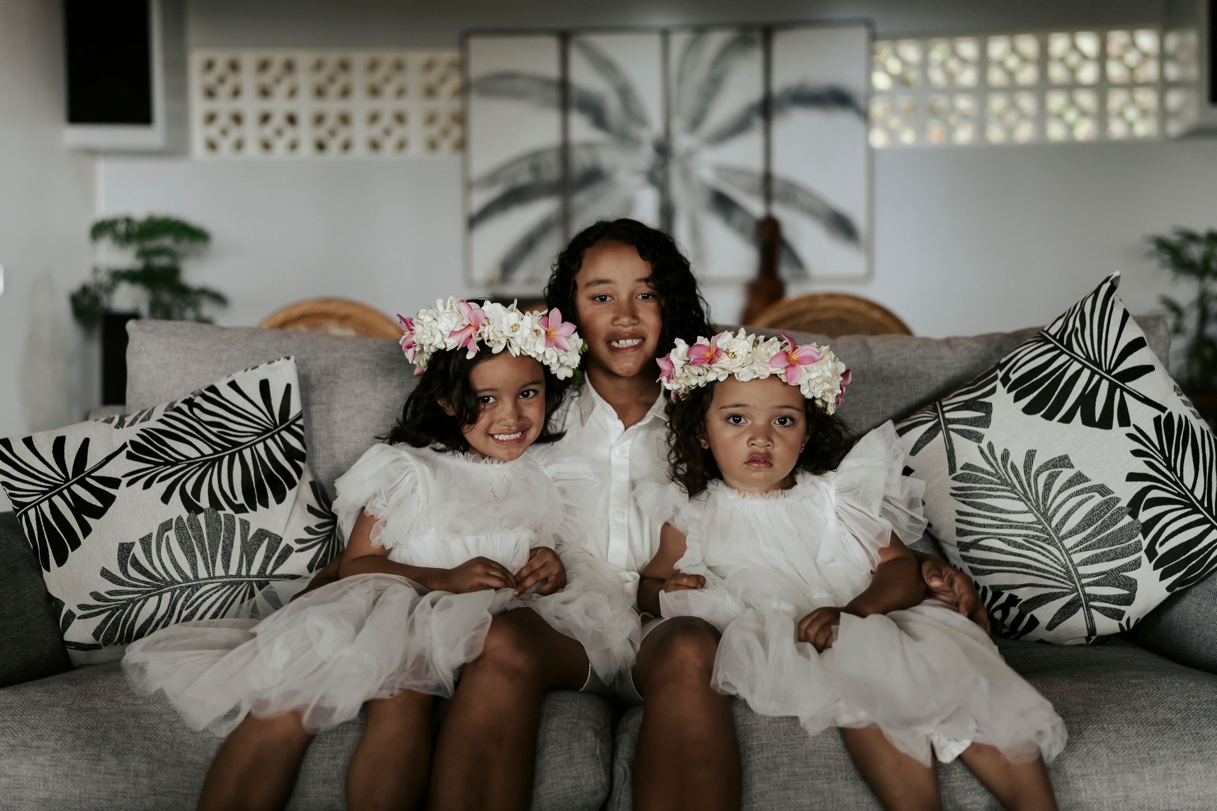 Two sisters with their brother on couch surrounded by tropical cushions – playful indoor family portrait in Rarotonga