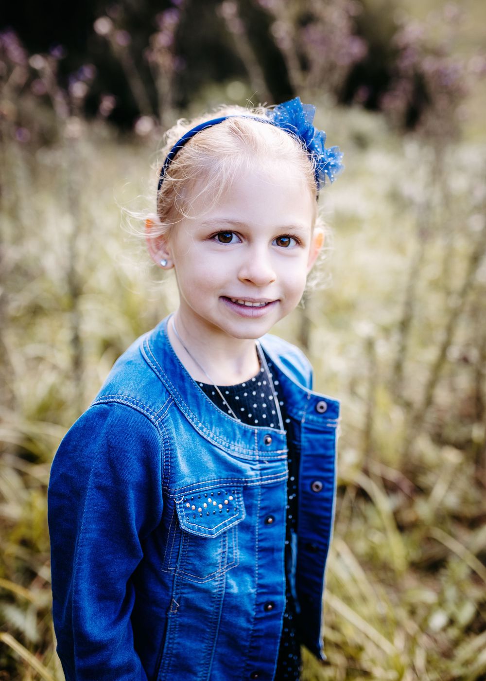 Young person in denim jacket with blue bow accessory poses outdoors in natural setting.