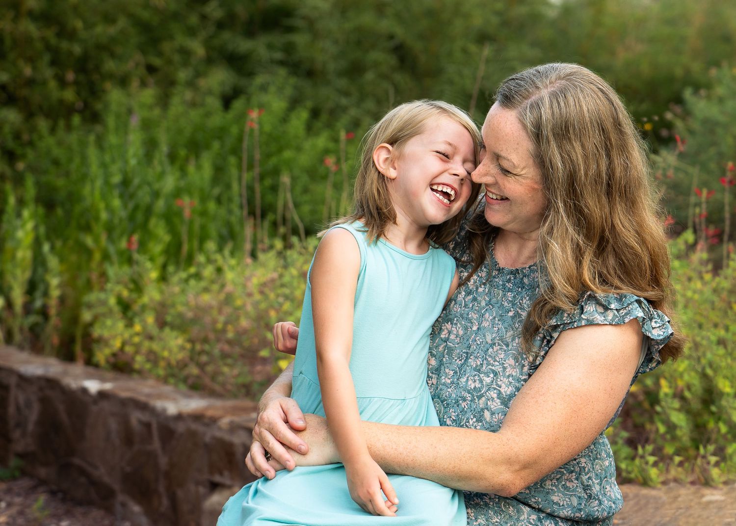a young girl in a blue sundress sitting on her mother's lap in Landa Gardens and they are both laughing