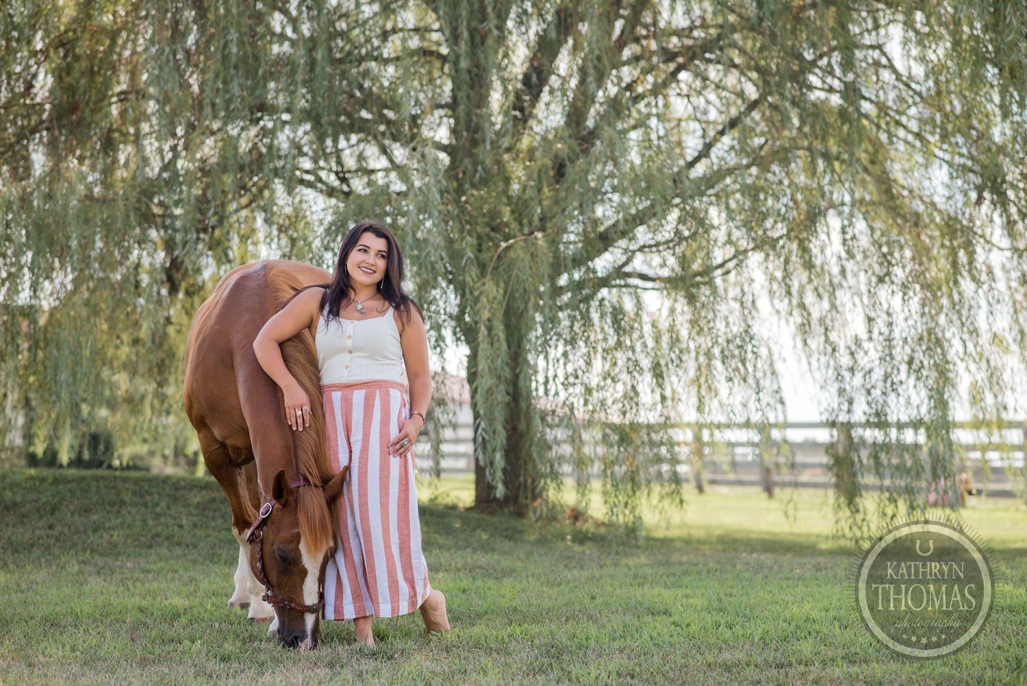 A person in striped dress stands beside grazing chestnut horse under willow trees.