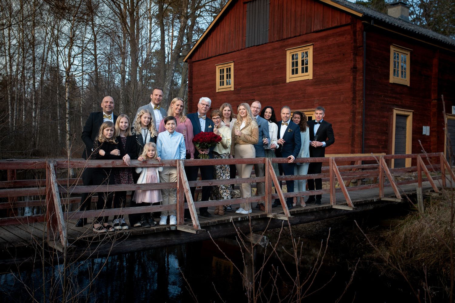 Wedding party gathered on wooden bridge in front of red Swedish cottage.