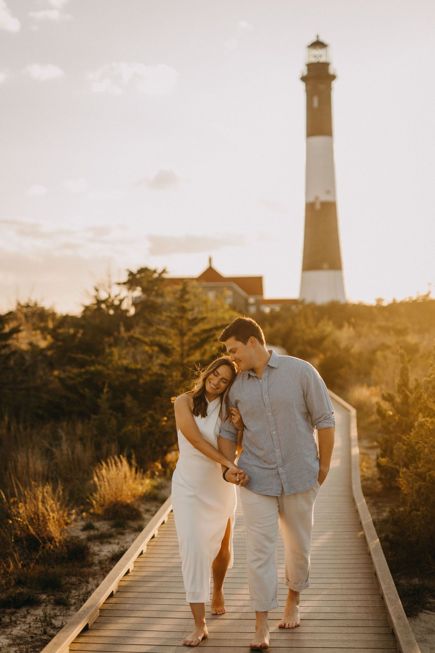 Couple walking along boardwalk path with lighthouse in background during golden hour.