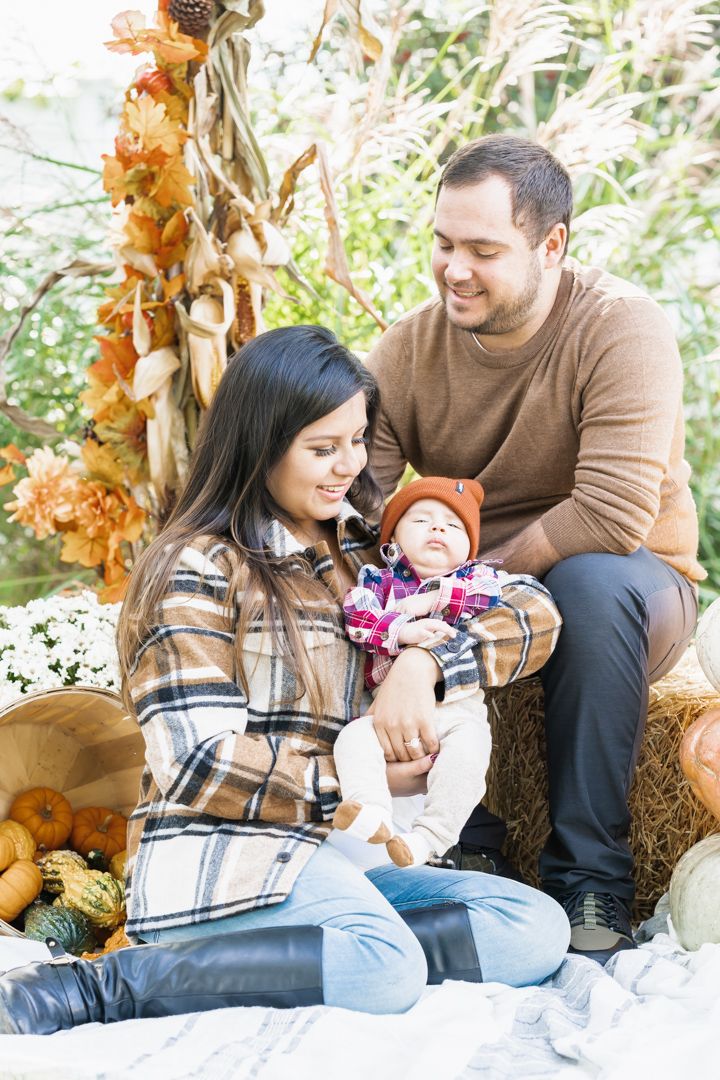 Family sits on blanket with baby among pumpkins and autumn decorations.
