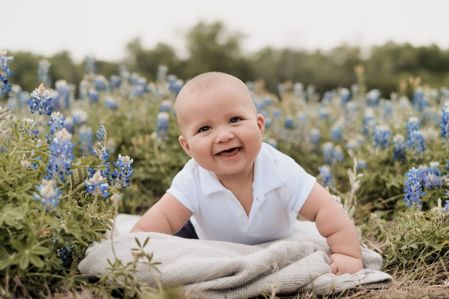 Baby in white outfit smiling among bluebonnets and blanket.