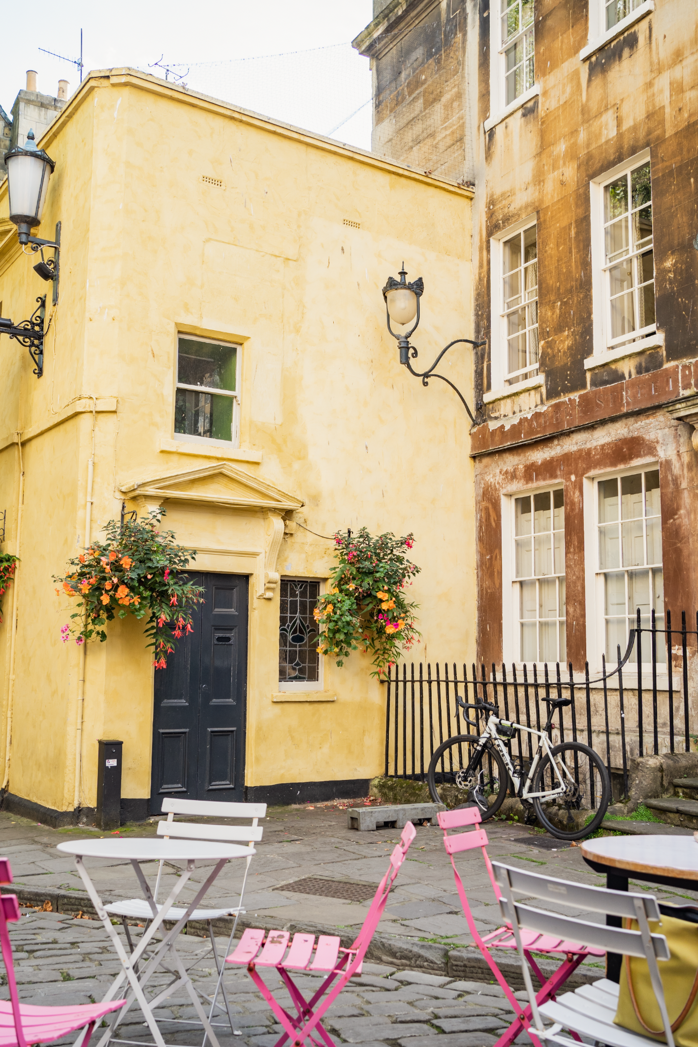 Yellow building with blue door, pink cafe chairs, and hanging flower baskets in charming European street.