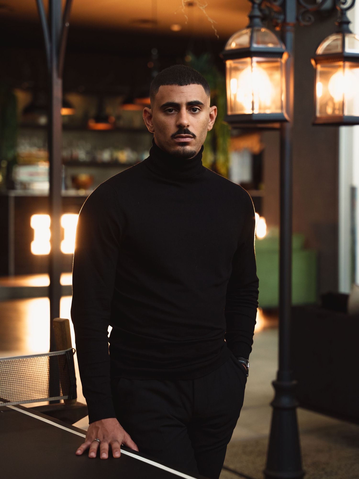 Moody portrait in black turtleneck against warm restaurant lighting with hanging lamps.