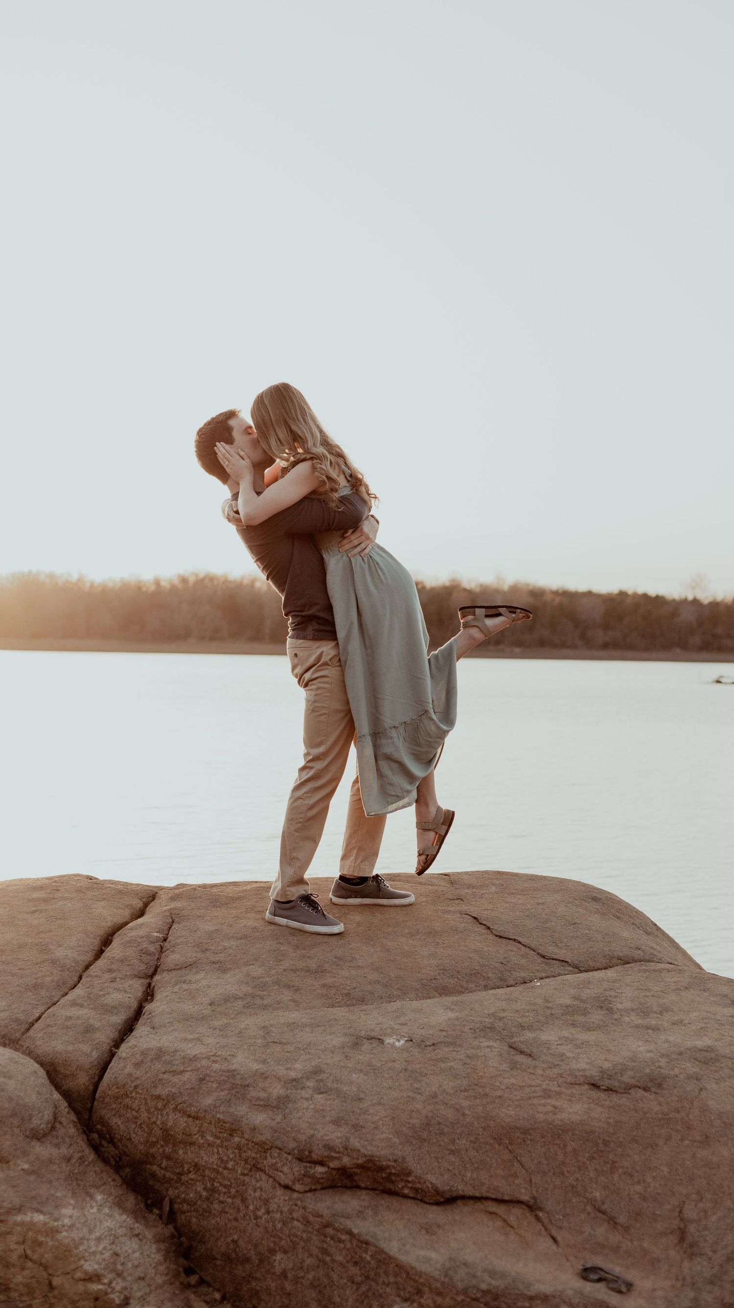 A couple embraces on a rocky cliff overlooking a serene lake at sunset.